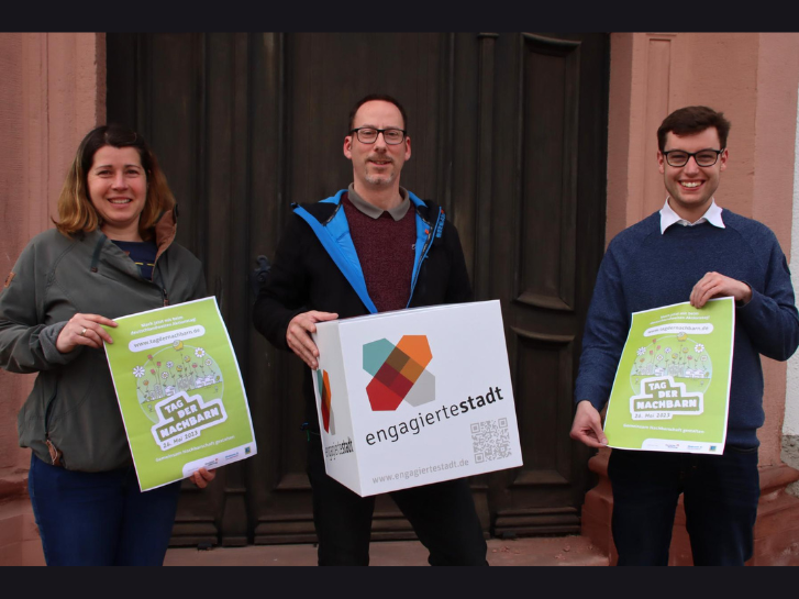 Auf dem Bild sind drei Personen abgebildet. Von links nach rechts Silke Reis, Stephan Germann und Marius Schmidt. Sie halten Flyer zum Tag der Nachbarn in den Händen und einen Würfel mit der Aufschrift Engagierte Stadt