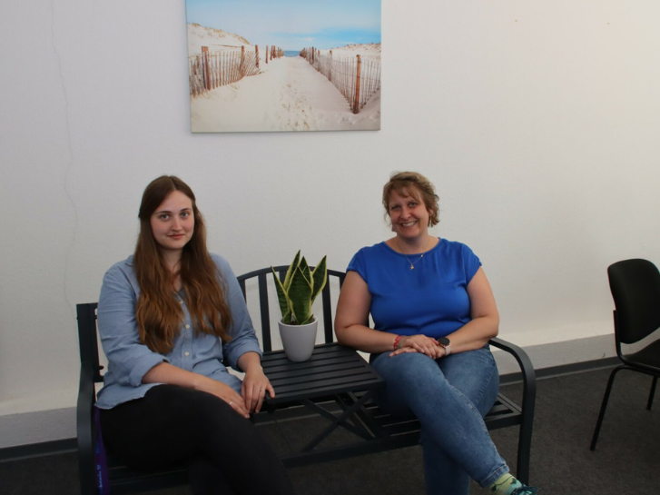 Franziska Wolff und Kerstin Biehal von der Gemeinwesenarbeit der Diakonie Bergstraße sitzen auf einer Bank, in ihrer Mitte steht eine Pflanze. An der Wand dahinter hängt ein Bild von einer Dünenlandschaft