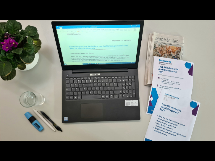 Auf einem Tisch steht ein Laptop mit einem Bewerbungsanschreiben. Daneben liegen Stifte, ein Glas Waser, eine Blume und Blätter plus eine Zeitung