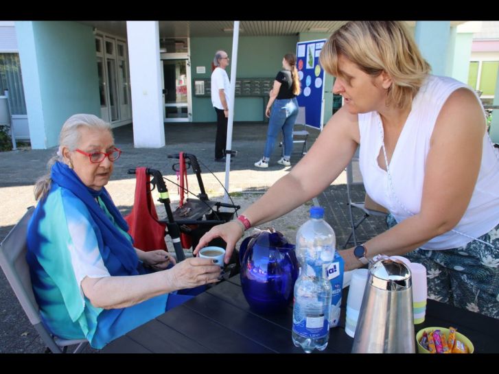 Kerstin Biehal reicht Frau Edith Bohn eine Tasse Kaffee. Im Hintergrund steht Franziska Wolff, die sich mit einem Mann unterhält.