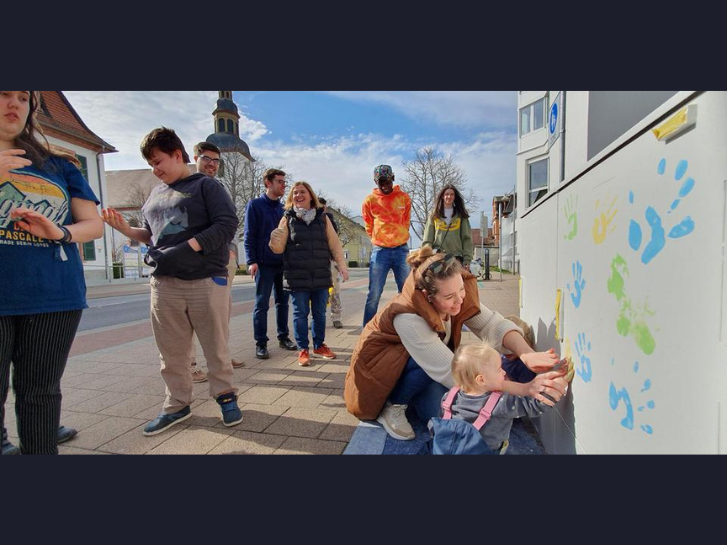 Eine Gruppe von Menschen, Kinder und Erwachsene stehen vor einem Stromkasten. Ein kleines Kind macht mit Hilfe einer Frau einen Handabdruck mit Farbe auf den Stromkasten