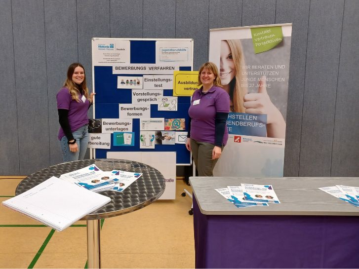 Auf dem Bild sind Janine Klingler und Kerstin Biehal aus der Fachstelle Jugendberufshilfe zu sehen. Sie stehen vor einer Pinnwand und einem Roll-Up der Fachstelle Jugendberufshilfe. Im Vordergrund steht ein Stehtisch und eine lila Messetheke.