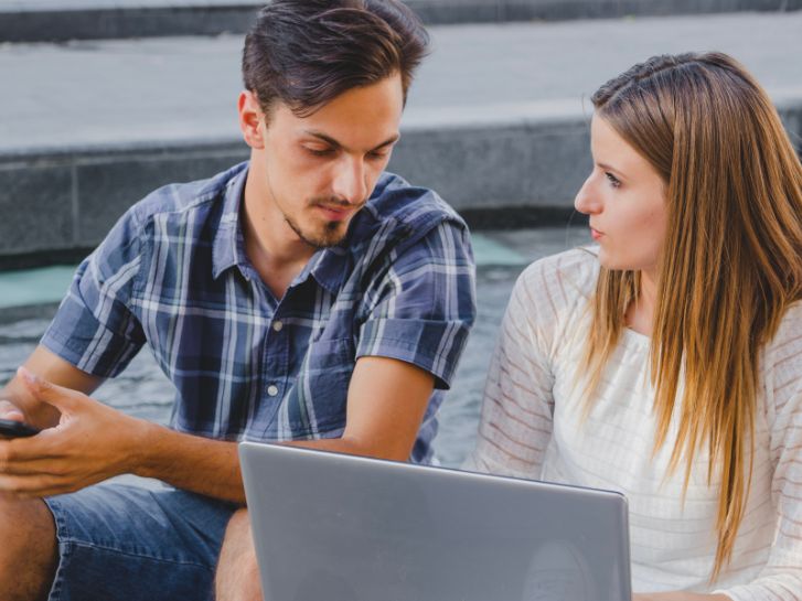 Ein Mann und eine Frau unterhalten sich. Die Frau hat einen Laptop in der Hand. Sie sitzen nebeneinander.