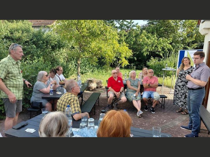 Mehrere Personen sitzen in einem Garten und hören einem Mann zu, der zu ihnen spricht. Zwei weitere Personen stehen.