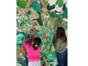 Zwei Kinder, die an dem Kunstprojekt vom BWF Rimbach teilnehmen, hängen von ihnen gestaltete Bilder an eine bunt gestaltete Wand, die einen Urwald darstellt.