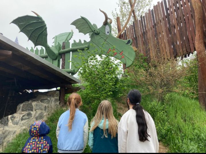 Vier Kinder schauen auf einen grünen Drachen aus Holz, der auf einem Hügel aufgebaut wurde.