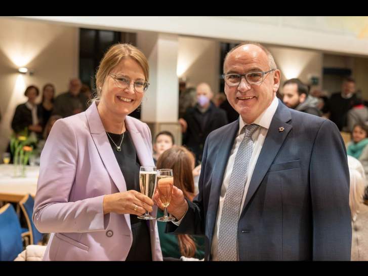 Dekanin Sonja Mattes und der scheidende Dekan Arno Kreh stoßen mit einem Sektglas an. Im Hintergrund sind die Gäste des Empfangs teilweise zu erkennen.