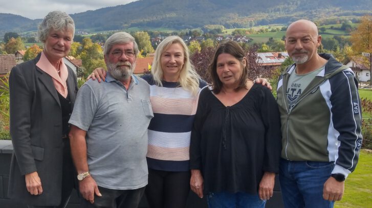 5 Personen, 2 Männer und 3 Frauen stehen draußen, im Hintergrund Umgebung und einige Häuser Landschaft von zu sehen