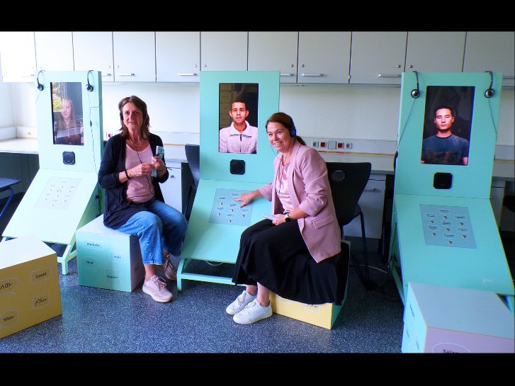 Marion Persson und Petra Popp sitzen neben einer Station der Ausstellung "Youniworth". Auf Bildschirmen sind Jugendliche zu erkennen, die von ihrer Fluchterfahrung berichten.