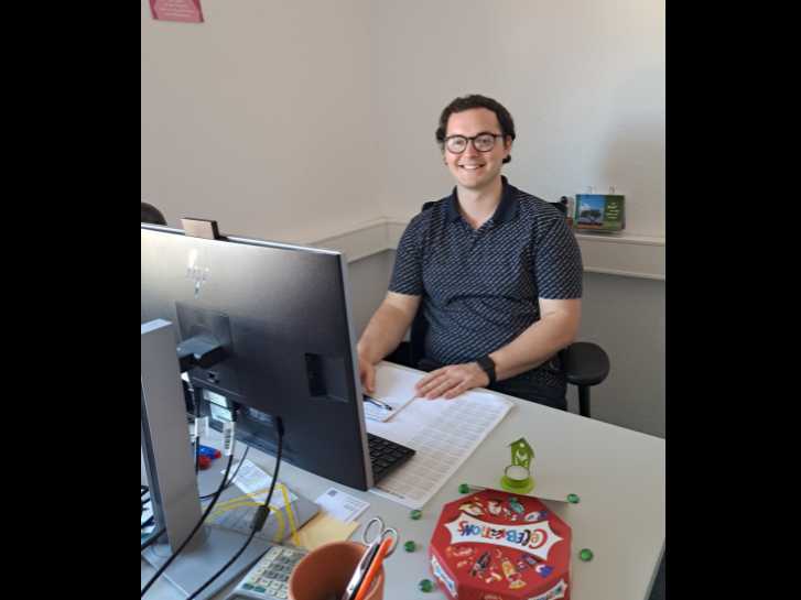 Hannes Schlauderer sitzt vor seinem PC im PSZ Rimbach und lächelt in die Kamera. Zur Begrüßung hatte ihm die Leitung des PSZ Rimbach eine Packung 'Celebrations' geschenkt, die vorne auf dem Schreibtisch steht.