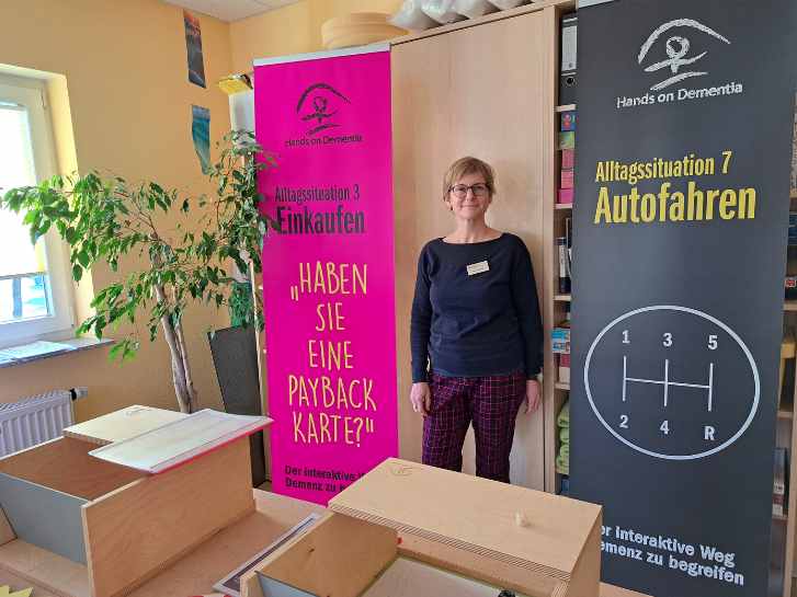 Im Psychosozialen Zentrum in Rimbach steht Frau Gürtler zwischen Rollups. Der pinke Rollup ist mit der Aufschrift "Alltagssituation Einkaufen" beschriftet. Darunter steht: Haben Sie eine Payback-Karte Auf dem linken, dunkelblauen Rollup steht: "Alltagssituation Autofahren". Im Vordergrund stehen auf Tischen angeordnet zwei Holzboxen, die Alltagssituationen simulieren sollen.