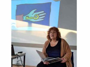 Felicia Schöner sitzt auf einem Stuhl mit dem aufgeschlagenen Buch in der Hand. Im Hintergrund das Bild einer Hand mit einem Schlüssel, das Schöner gemalt hat.