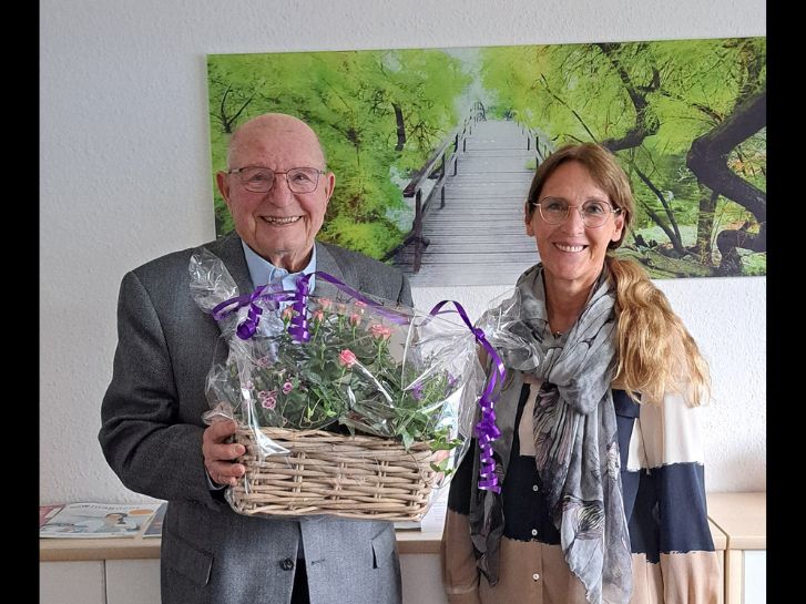 Vor einem Bild mit einer Holzbrücke, umgeben von hellgrün belaubten Bäumen stehen Rudi Jakob links und Susanne Hagen. Rudi Jakob hält einen Korb mit einem Blumengesteck in der Hand.