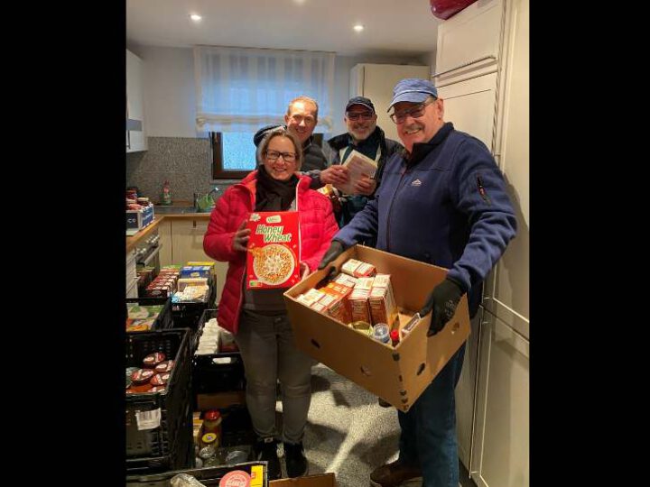 Drei Männer und eine Frau stehen in der Küche von Familie Olf. Der Mann im Vordergrund präsentiert einen Karton mit Lebensmitteln.