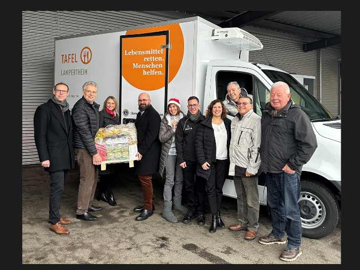 Im Autohaus in Worms stehen Vertreterinnen und Vertreter vor einem Kühlauto, das für die Tafel Lampertheim bestimmt ist. Zwei Personen halten einer Kiste mit Lebensmitteln.