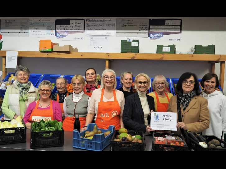 Bei der Tafelausgabestelle in Rimbach stehen 12 strahlende Frauen vor Kisten mit Tomaten, Salat, gemischtem Gemüse und Obst. Zwei Frauen halten einen "Scheck" über 2000 Euro. Im Hintergrund befinden sich Regale mit blauen Gemüsekisten. Darüber sind weitere Spendenübergabe-Schecks wie Poster an der Wand befestigt.