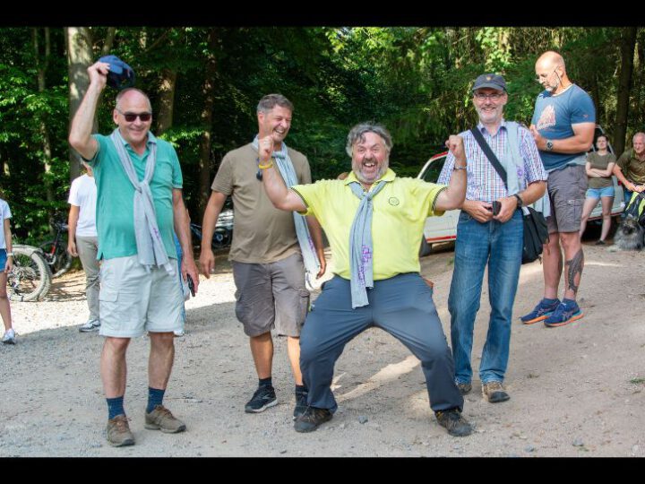 Pfarrer Ningel (Mitte) reißt freudig die Arme hoch. Die Wette anlässlich des Tafel-Aktionstages wurde gewonnen. Von l. Dekan Arno Kreh, Tobias Lauer (Leiter Regionale Diakonie Bergstraße) , Probst Stephan Arras. Sie stehen auf dem Kiesplatz auf der Tromm. Im Hintergrund Gäste der Veranstaltung.