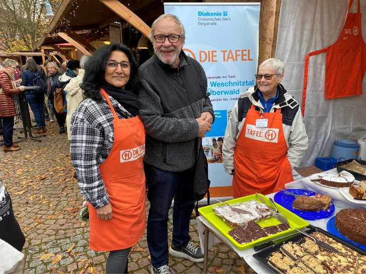 Drei Tafelmitarbeiter, 2 Frauen und 1 Mann stehen lächelnd hinter dem Tisch, auf dem Bleche und Kuchenplatten mit selbstgebackenem Kuchen stehen.