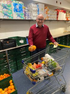 Dekan Arno Kreh schiebt einen Einkaufswagen und hält zwei Orangen in der Hand. Im Hintergrund befinden sich auf Regalen und in grünen Plastikkisten die Waren die bei der Tafel Rimbach an die bedürftigen Menschen ausgegeben werden 