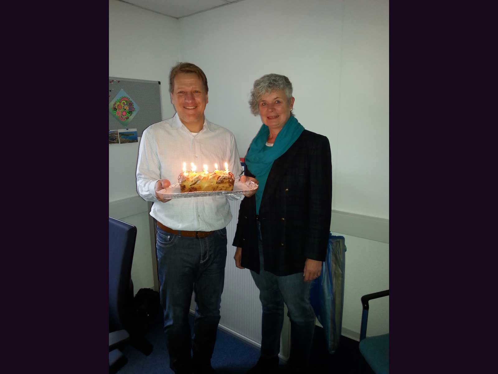 Zwei lachende Sozialpädagogen mit einem Kuchen auf dem zehn Kerzen scheinen.