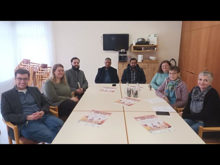 Acht Personen sitzen an einem Tisch und machen WErbung für die Aktion Warmes Essen für Alle in Lampertheim