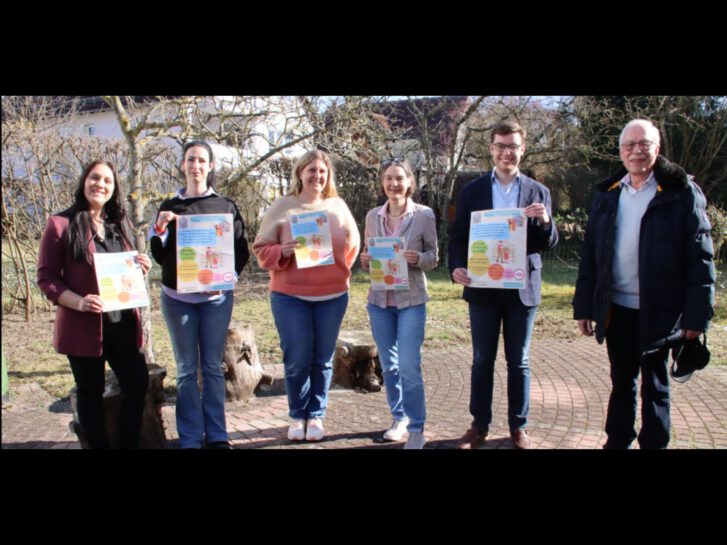 In einem Garten stehen sechs Personen nebeneinander und halten ein Schild in die Höhe mit Werbung für die Veranstaltung Wohnen für Hilfe