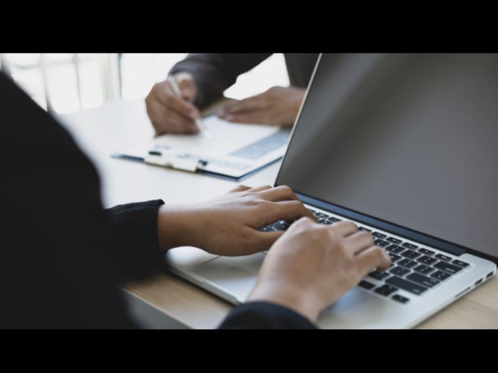 Eine Person tippt auf einem Laptop. gegenüber schreibt jemand auf einem Klemmbrett.