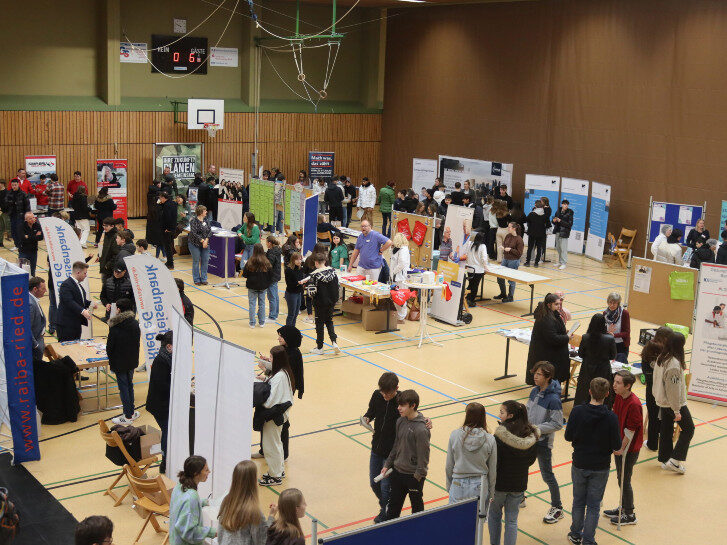 Auf dem Bild sieht man mehrere Aussteller, u.a. Betriebe sowie Schülerinnen und Schüler in einer Turnhalle. Sie besuchen die Berufsinformationsbörse der Erich Kästner Schule in Bürstadt