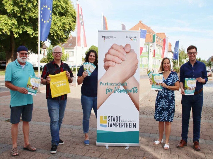 Auf dem Bild sind fünf Personen zu sehen. In der Mitte steht ein Role Up der Partnerschaft für Demokratie und mehrere Personen halten Flyer und Zettel hoch.