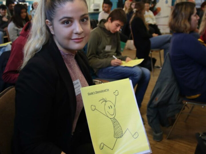 Eine junge Frau hält ein gelbes Blatt in der Hand auf dem ein Menschen gemalt ist mit der Beschriftung Guter Demokrat. Im Hintergrund sitzen viele Menschen auf Stühlen