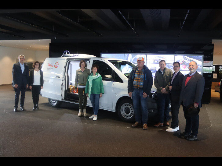 8 Personen stehen vor einem neuen weißen Kühlwagen der Tafel in einem Ausstellungsraum.