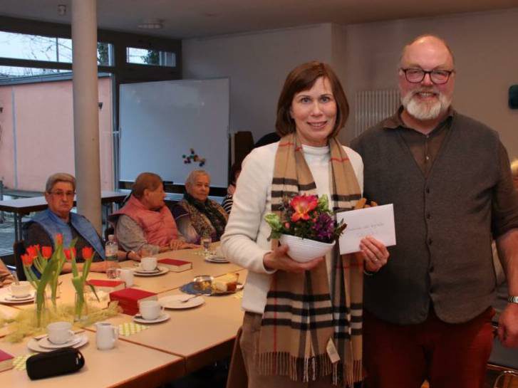 Die Tafelkoordinatorin steht neben dem Pfarrer und hält den Scheck und ein Blumengesteck. Sie befinden sich im Seniorenzentrum. Hinter ihnen befindet sich die Kaffeetafel, um die mehrere Senioren sitzen.