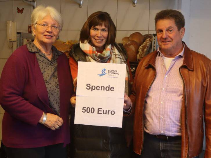 Drei Personen stehen im Tafelgebäude. Die Person in der Mittel hält ein Plakat mit dem Text "Spende 500 Euro".